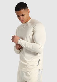 Maglietta sportiva a maniche lunghe di colore crema con un tessuto testurizzato, dotata di scollatura arrotondata e logo stampato sul lato sinistro.