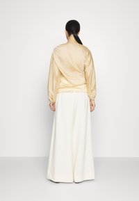 Vaaleanruskea satiininen puffihihainen paita yhdistettynä valkoisiin leveälahkeisiin housuihin. Sileä pinta, yksinkertainen muotoilu ja minimimäärä näkyvää yksityiskohtaa takaa.