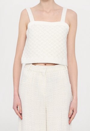 Kvinde iført en hvid tekstureret ærmeløs crop top og matchende højtaljede, vidde ben bukser mod en ensfarvet baggrund.