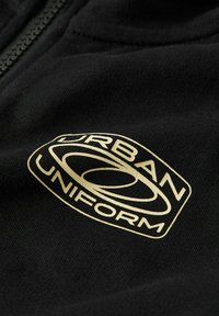 Tissu noir avec un logo "Urban Uniform" clair et un design ovale stylisé, à côté d'un détail de fermeture éclair noir.