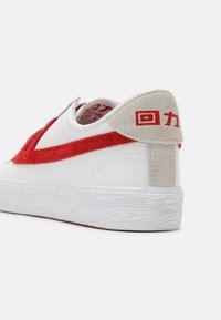 Bijele tenisice s crvenim suede detaljima, zaobljenog oblika, s teksturiranom gumenom potplatom i teksturiranim bež petom koja ima grafički detalj.