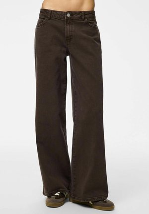 Persoon draagt losse donkerbruine jeans met wijde pijpen en bruine casual schoenen, zichtbaar van de taille tot de voeten tegen een egale achtergrond.