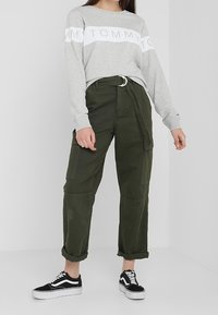 Szara bluza z białym paskiem z logo, oliwkowo-zielone spodnie cargo z podwiniętymi nogawkami oraz czarno-białe sneakersy z kontrastowymi sznurowadłami.