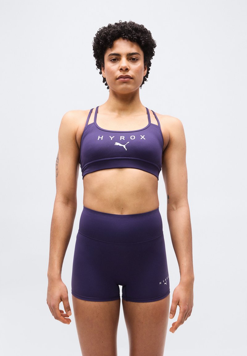 Αθλητικός άνθρωπος με κοντά σγουρά μαλλιά που φοράει μωβ αθλητικό σουτιέν HYROX Puma και ταιριαστά σορτς, στέκεται μπροστά σε ανοιχτό γκρι φόντο.