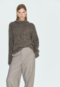 Bruine gebreide trui met een hoge kraag, voorzien van een textuurpatroon. Gecombineerd met lichtgrijze, loszittende broeken.