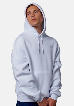 Joven con sudadera blanca con capucha puesta, mirando hacia un lado, con las manos juntas al frente, sobre un fondo liso.
