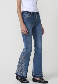 Jeans de mezclilla azules acampanados con coloridos bordados en los puños, con un diseño clásico de cinco bolsillos y un sutil desgastado.
