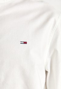 Biela bavlnená košeľa s golierom na gombík, na ľavej hrudi s malým vyšitým logom v modrej, červenej a bielej farbe.