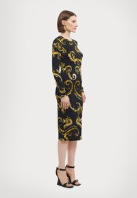 Svart langermet kjole med gule virvelmønstre og tekst; tettsittende silhuett; kne-lengde. Kombinert med svarte høyhælte sandaler.