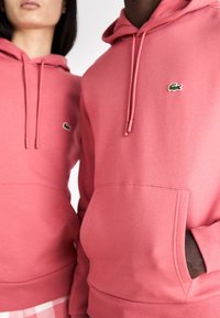 Deux personnes portant des sweats à capuche Lacoste roses, l'une avec la main dans la poche avant, toutes deux affichant le logo crocodile brodé.