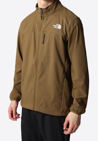 Brun softshell-jakke med høj krave, lynlåslukning foran og sidelommer. Designet har elastiske manchetter og en lille logo på brystet.