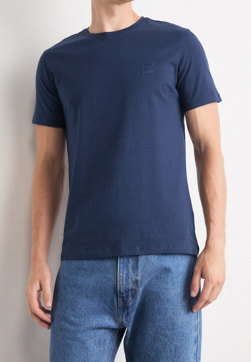 Homme portant un t-shirt uni bleu marine à manches courtes et un jean en denim bleu clair sur un fond uni.