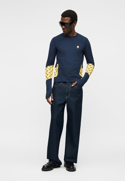 Άνδρας που φοράει μπλε σκούρο μακρυμάνικο πουκάμισο με κίτρινες σχέδια σε σχήμα ημισελήνου στα μανίκια και τις πλευρές, σκούρο τζιν, μαύρα παπούτσια και μεγάλα μαύρα γυαλιά ηλίου.