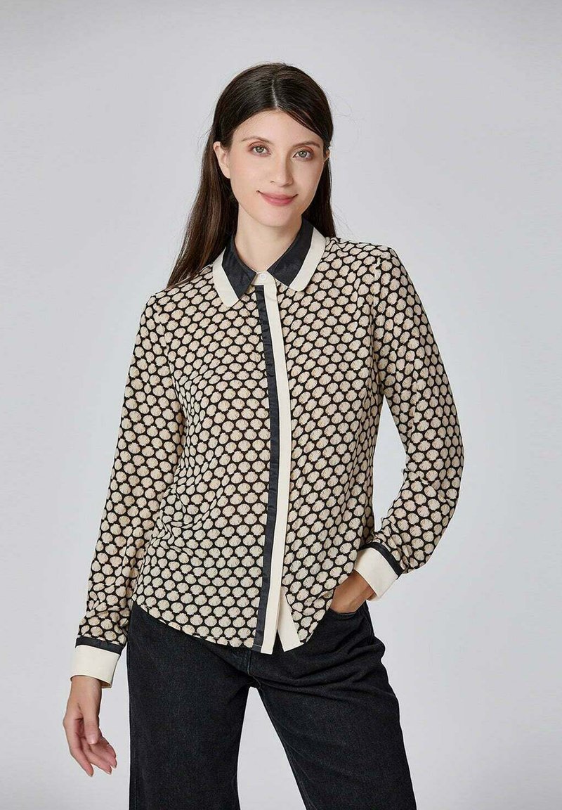 Blouse noire et crème avec une texture à motifs, à manches longues et un col noir ; présente un devant à boutons et des poignets contrastants.
