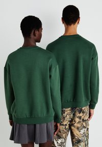 Felpe verdi in tessuto felpato, a vestibilità comoda, con spalle scese, polsini e orli a coste. Un modello indossa una gonna grigia, l'altro dei pantaloni a fantasia.