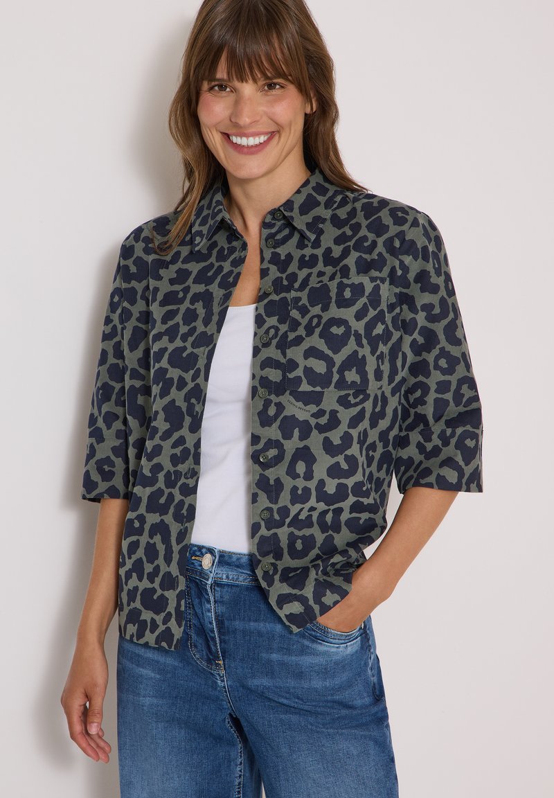 Femme souriante portant une chemise à boutons imprimée léopard verte et noire sur un haut blanc et un jean bleu, avec une main dans la poche.