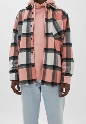 Personne portant un sweat à capuche rose sous une chemise à carreaux rose, noire et blanche boutonnée, et un jean bleu clair sur un fond uni.