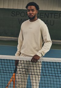 Cream sweatshirt and joggers with ribbed cuffs, displaying a logo. Standing by a tennis net, holding an orange racket on a blue court.