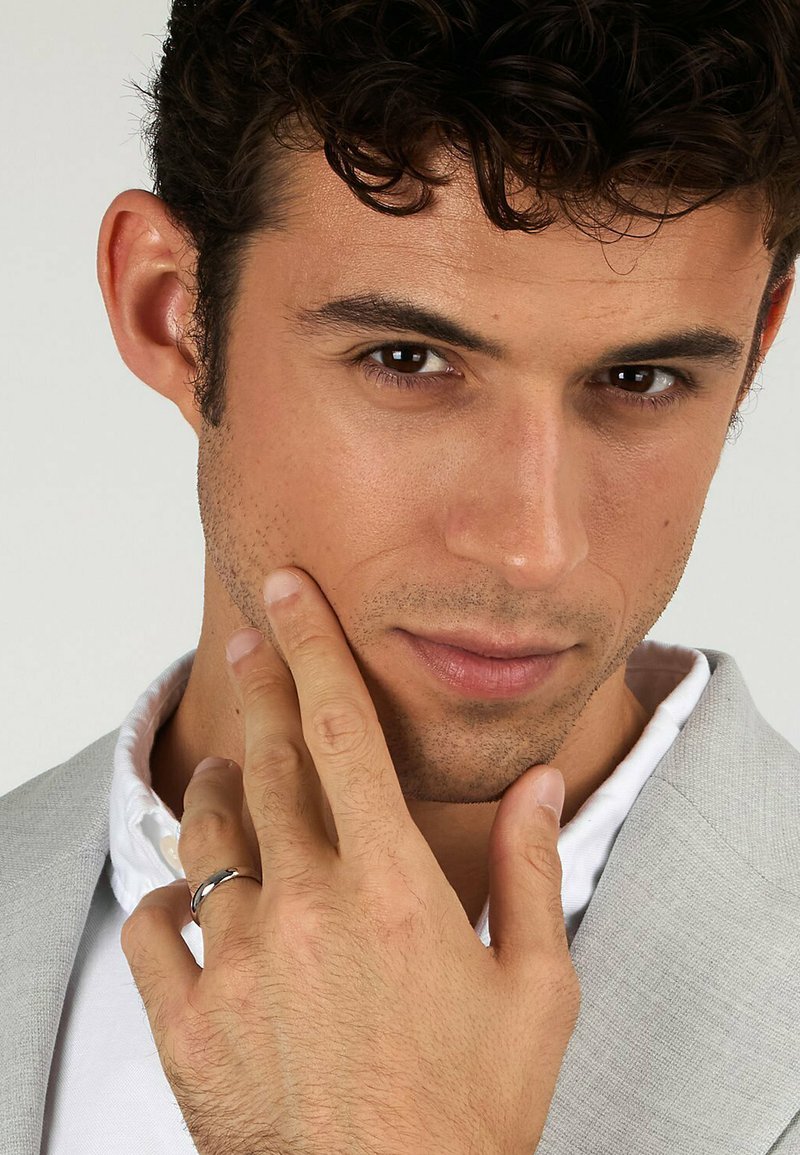 Silvring på vänster hand; ljusgrå kostymjacka, vit skjorta. Kort, lockigt mörkt hår och rakad ansikte med neutral uttryck.