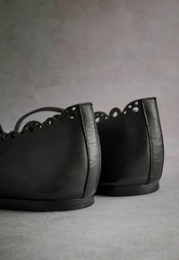 Ballerine in pelle nera con bordo ondulato e dettagli perforati; suola piatta, texture liscia e design della parte posteriore arrotondata.