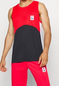 Chemise sans manches rouge et noire avec encolure ronde, écusson logo Nike, et panneaux latéraux contrastants ; assortie à un pantalon rouge coordonné.