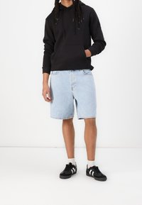 Felpa nera con tasca frontale, pantaloni corti in denim blu chiaro e sneakers nere con strisce bianche. Texture liscia e vestibilità casual.
