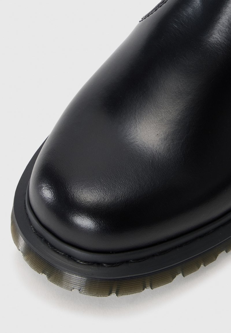 Botte en cuir noir avec une surface lisse et brillante, un bout rond, une semelle en caoutchouc texturée contrastante et des coutures visibles le long du bord.