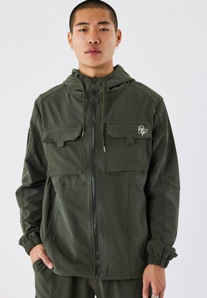 Olijfgroene jas van gladde stof, met een rits aan de voorkant, twee borstzakken, elastische manchetten en een capuchon met trekkoord.