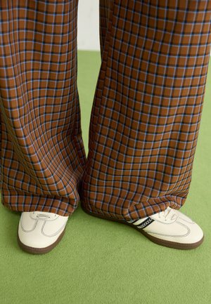Bruine en blauwe geruite wijde broek, gecombineerd met witte schoenen met zwarte accenten en een gestructureerde zool, gezet tegen een groene achtergrond.