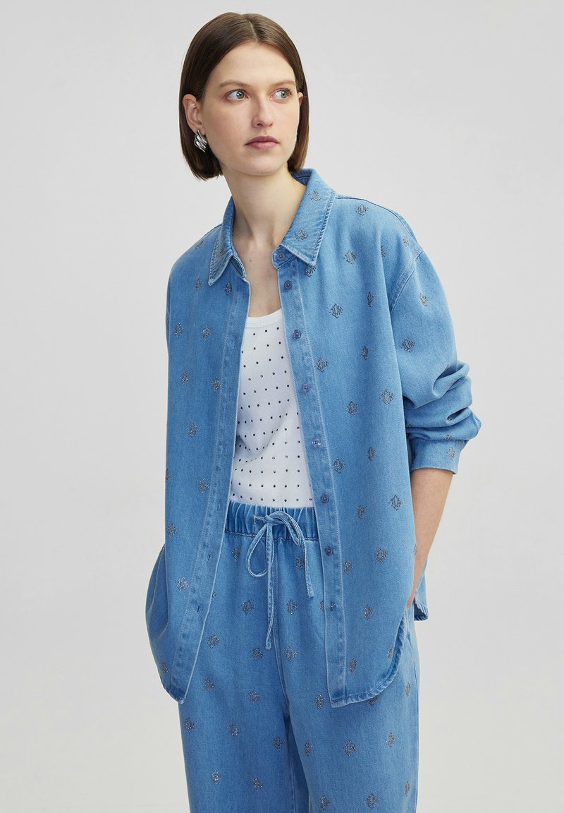 Camicia di jeans azzurro chiaro con maniche lunghe, bottoni a pressione e fantasie floreali ricamate. Indossata su un top a canotta bianco con pois.