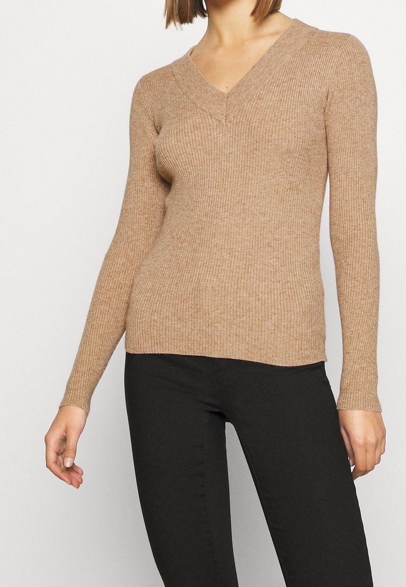 Pull en maille côtelée beige avec col en V, manches longues et silhouette ajustée, associé à un pantalon noir moulant, mettant en valeur la texture et le design.