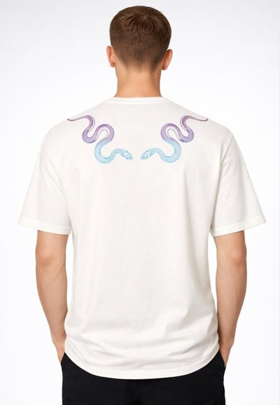 Uomo che indossa una t-shirt bianca con due serpenti simmetrici blu e viola stampati sulla parte superiore della schiena, in piedi con le mani in tasca.