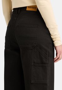Pantalones cargo negros con múltiples bolsillos, hechos de tejido duradero. Presentan una etiqueta de Timberland en la cintura y un diseño de pierna recta.