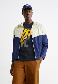 Nike vēja jakas viegli dzeltenā un tumši zilā krāsā ar kapuci un rāvējslēdzēju. Pāris ar melnu T-kreklu ar dzeltenu tekstu un brūnām biksēm.