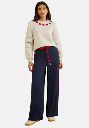 Naine, kes kannab helehalli kampsunit punaste südamekujuliste detailidega krae ääres, tumepunaste triipudega laia lõikega tumesiniseid pükse ja beeže jalanõusid.