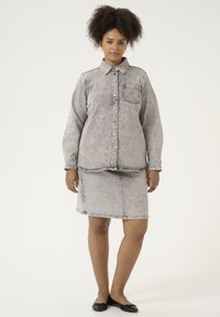 Lys grå denim skjorte med frontlomme og knapper, parret med en matchende tætsiddende nederdel. Flade sorte sko fuldender outfittet.