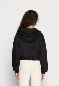 Sudadera negra corta con una gran capucha, hombros caídos y dobladillo elástico. Tejido suave con un ajuste relajado, combinada con pantalones claros.