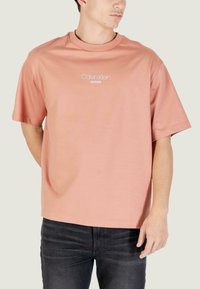 Camiseta de algodón rosa con mangas cortas y cuello redondo, con el logo "Calvin Klein JEANS" en blanco en el pecho. Combinada con vaqueros oscuros.