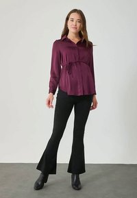 Γυναίκα φορεί μια μακριά μανίκια μωβ μπλούζα εγκυμοσύνης με ζωνάκι στη μέση, μαύρα καμπάνα παντελόνια και μαύρες μποτίνες μέχρι τον αστράγαλο, στέκοντας σε γκρι πάτωμα.