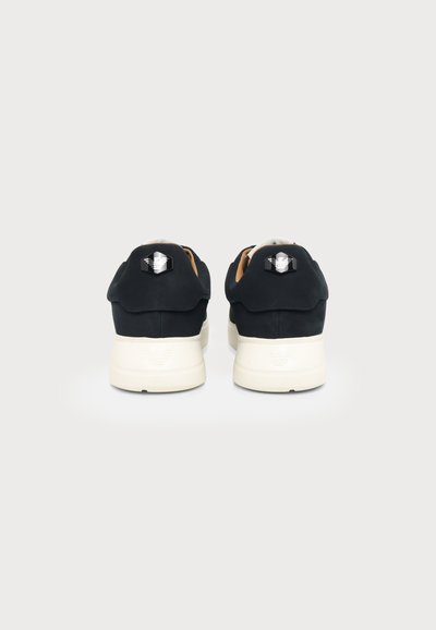 Paire de baskets noires avec semelles blanches visibles de dos, affichant un logo argenté réfléchissant sur la languette du talon.