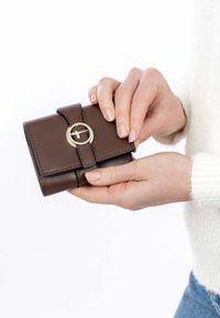 Rudas odinis piniginė su auksiniu apvaliu užsegimu, turinti lygią tekstūrą ir siuvinėtus akcentus, laikoma rankoje.