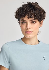 Ljusblå t-shirt med kort ärm, tillverkad av mjukt tyg, med en liten mörk logotyp på bröstet, rund halsringning och avslappnad passform.