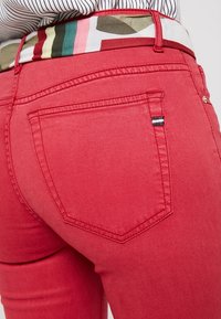 Jean rouge au design ajusté, avec des poches arrière et une ceinture à motifs contrastants dans des couleurs comme le vert, le rose et le beige.