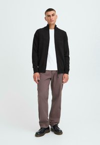 Sort zip-up cardigan over en hvid T-shirt, parret med lysbrune bukser og sorte sko. Glat stof, standard pasform, afslappet stil.