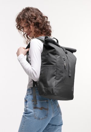 Giovane donna con capelli ricci che indossa una camicia bianca a maniche lunghe e jeans blu, che porta un grande zaino nero con chiusura a rotolo.