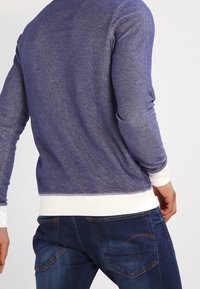 Námořnicky modrý pletený svetr s texturovaným povrchem, bílými žebrovanými manžetami a lemem, doplněný tmavě modrými džínami s šikmými zadními kapsami.