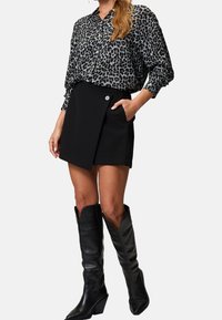 Schwarzer Wickelrock mit Seitentasche und Knopfdetail, kombiniert mit einer locker sitzenden grauen Bluse im Leopardenmuster und kniehohen schwarzen Lederstiefeln.
