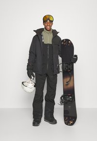Snowboarder wearing a black jacket, green mid-layer, black pants, and gloves, holding a snowboard and white helmet. Honeycomb goggles.