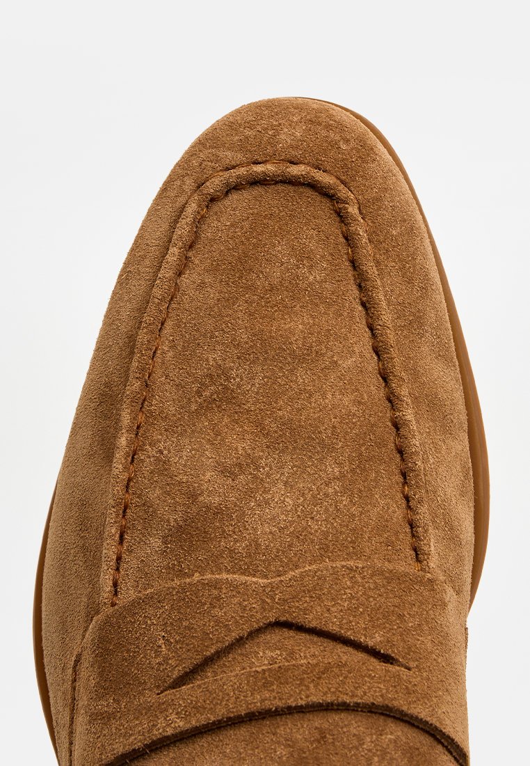 Primer plano de una loafer de ante marrón con costuras visibles y un detalle de tira elegante en la parte superior.