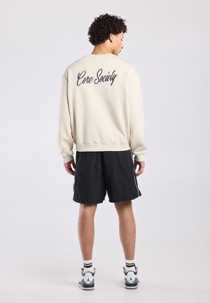 Jonge man met krullend haar staat met zijn rug naar de camera toe, draagt een crèmekleurige sweatshirt met de tekst "Core Society" en zwarte korte broek.
